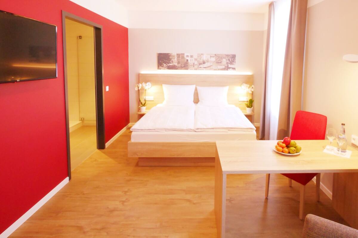 A hotel room with a bed, a desk with a chair, a table with fruit, and a TV on the wall.