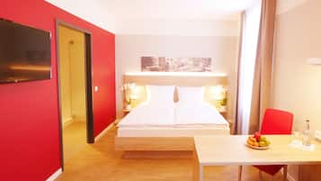 Ein Hotelzimmer mit einem Bett, einem Schreibtisch mit Stuhl, einem Tisch mit Früchten und einem Fernseher an der Wand.