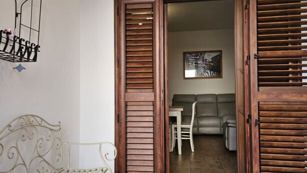Een kamer met een houten deur, een witte metalen bank, een grijze bank en een kleine tafel.