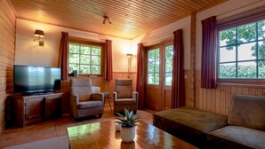 A cozy living room with a wooden ceiling, a flat-screen TV, two armchairs, a sofa, a coffee table with a book, and large windows with curtains. - Cozy Swedish group home in forest with private parking (Soest)
