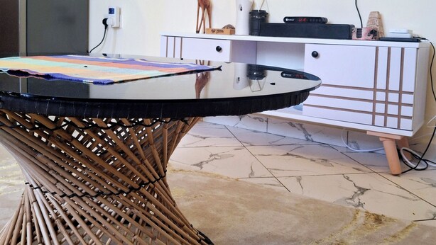 A modern living room with a flat-screen TV mounted on the wall displaying various apps. There is a round table with a woven base in the foreground, and a white cabinet with a TV on top in the background. - Zai Homes Watamu (Watamu)