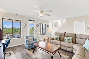 A living room with a sofa, armchair, coffee table, and a ceiling fan. There are windows with blinds, a clock, and a sign on the wall.