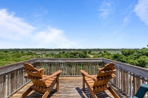 A wooden deck with two Adirondack chairs facing a scenic view of a lake and forest. - Shell Haven-Four Decks, Views, Pet friendly, Walk to beach! (Holden Beach)