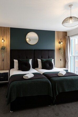 Uma cama de casal com roupa de cama verde escura, um espelho redondo acima e duas luminárias fixadas na parede.
