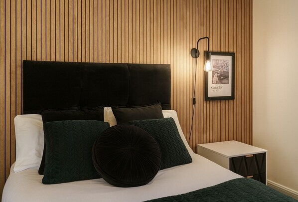 A bed with a black headboard, white pillows, and green and black pillows. There is a framed picture on the wall and a small bedside table with a lamp.