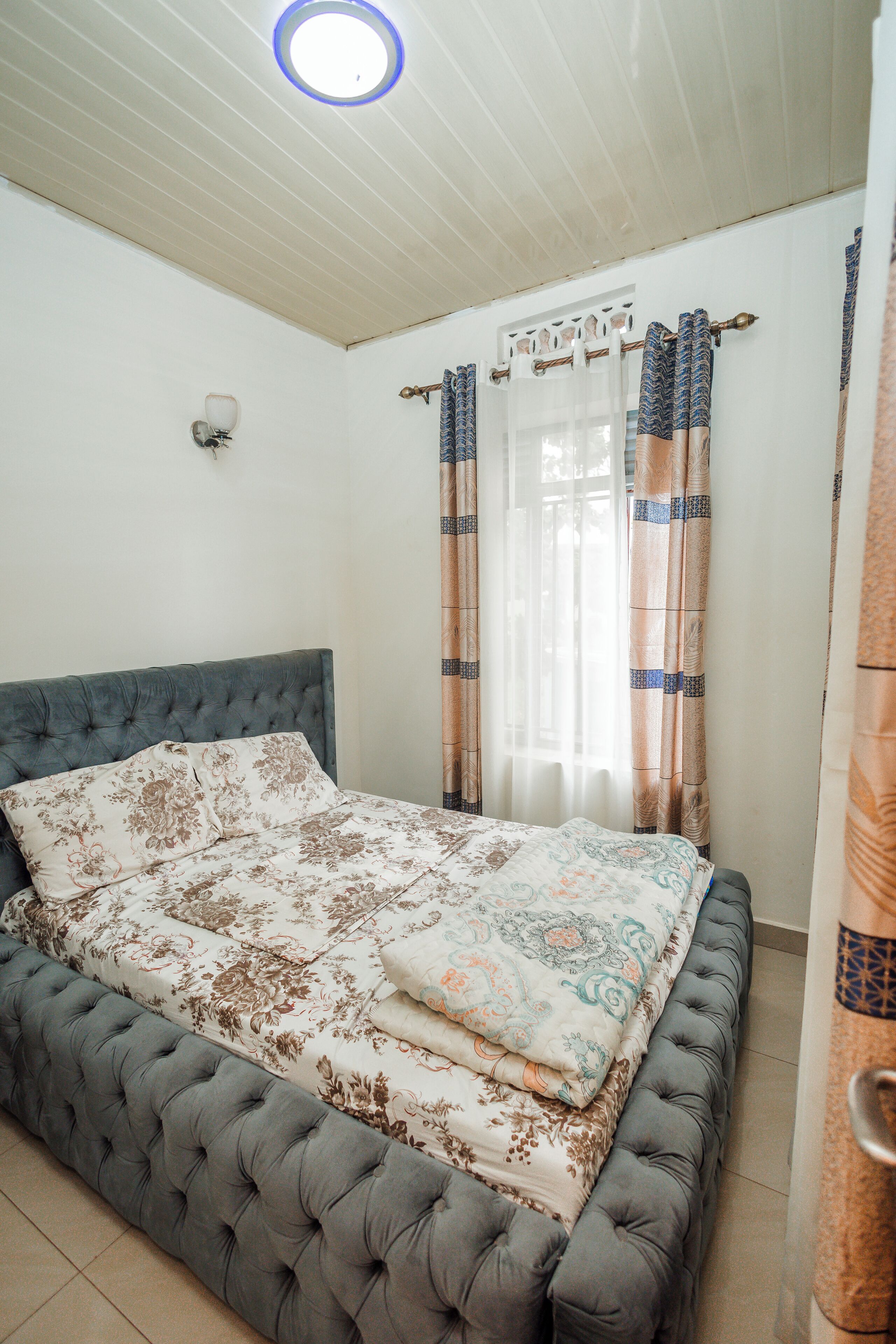 A bedroom with a tufted headboard, patterned bedding, and a window with curtains.