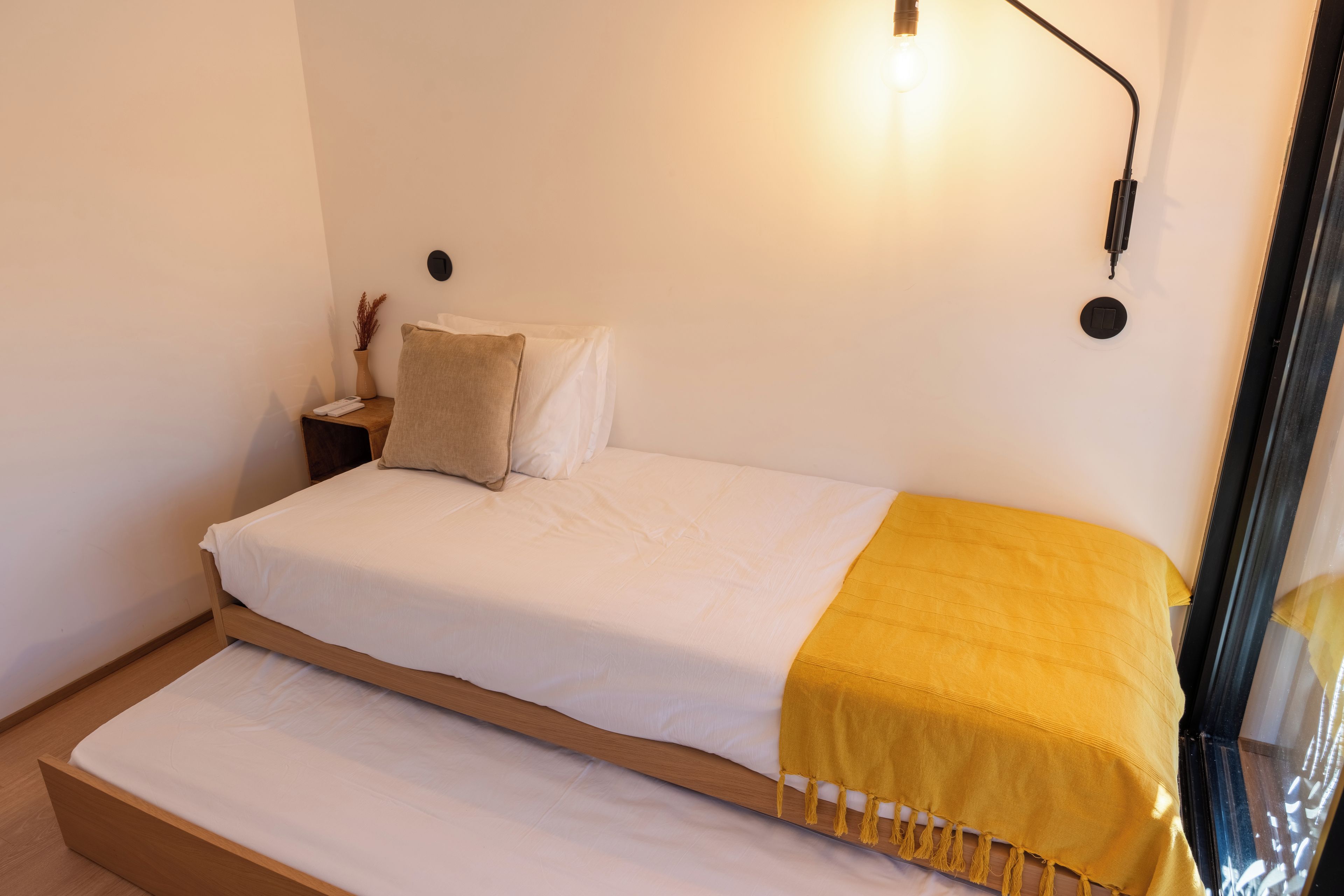 Una cama individual con colchón blanco y manta amarilla, una mesita de noche con un florero, y una lámpara de pared.