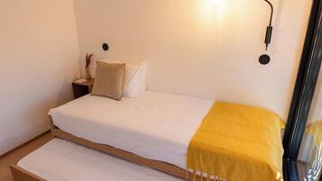 Una cama individual con colchón blanco y manta amarilla, una mesita de noche con un florero, y una lámpara de pared.