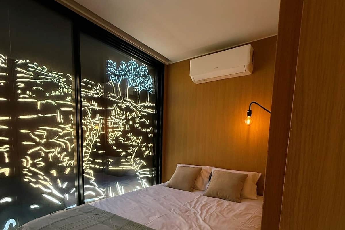 Un dormitorio con una cama grande, una mesita de noche, un aire acondicionado de pared y una ventana con luces.