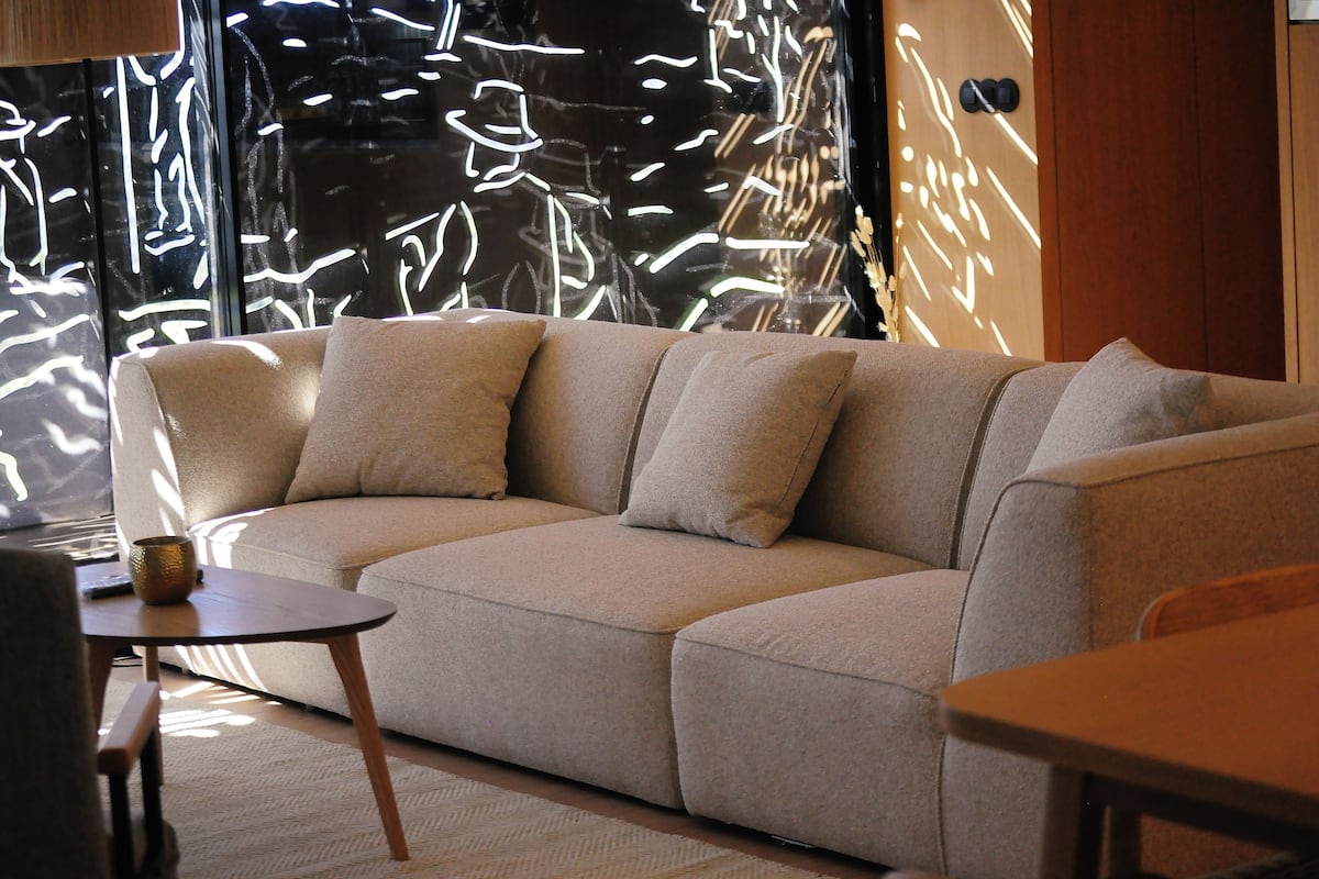 Un sofá seccional color beige con una pequeña mesa redonda y una silla en una sala de estar moderna con un ventanal grande y arte abstracto en la pared.