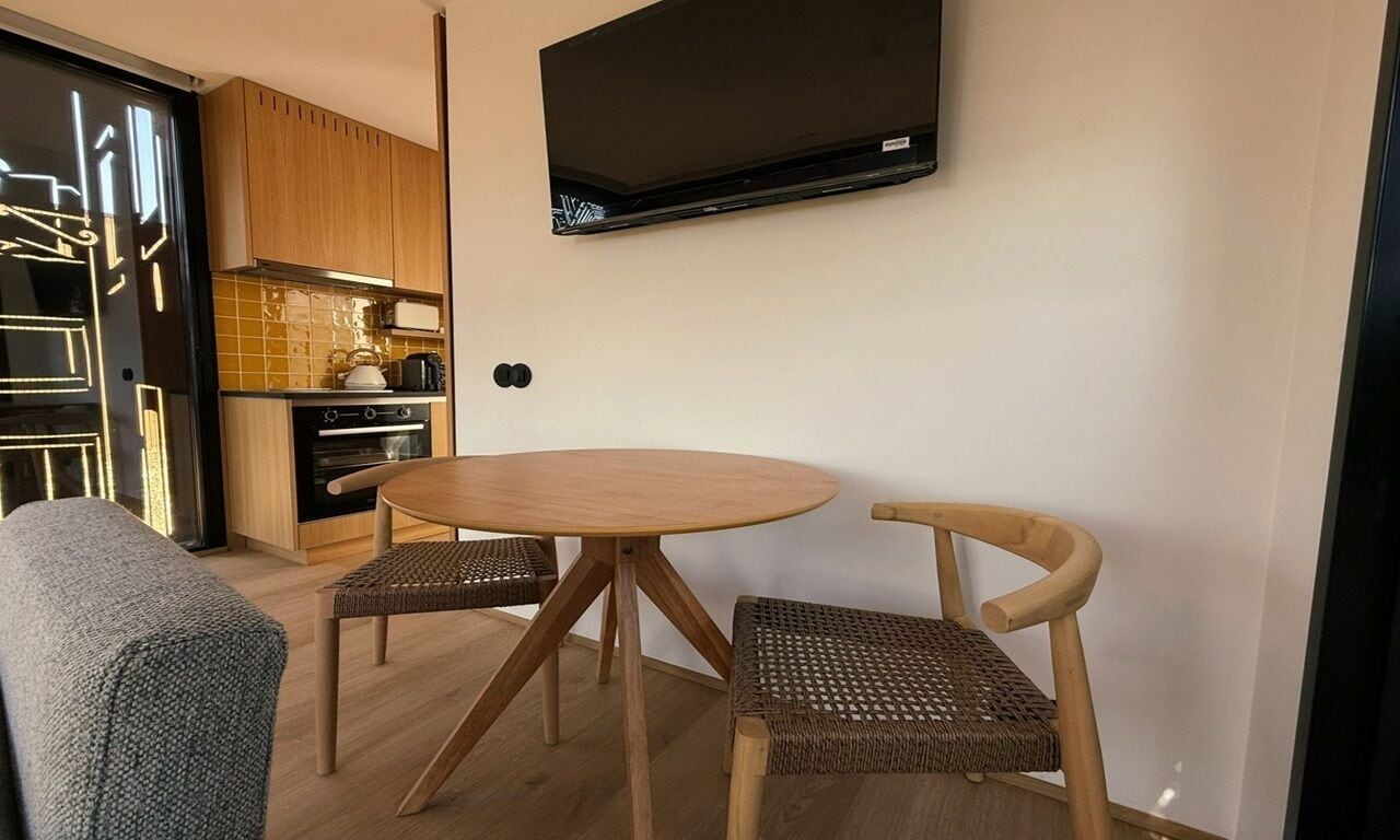 A compact kitchen with a TV mounted above a wooden dining table and two wicker chairs.