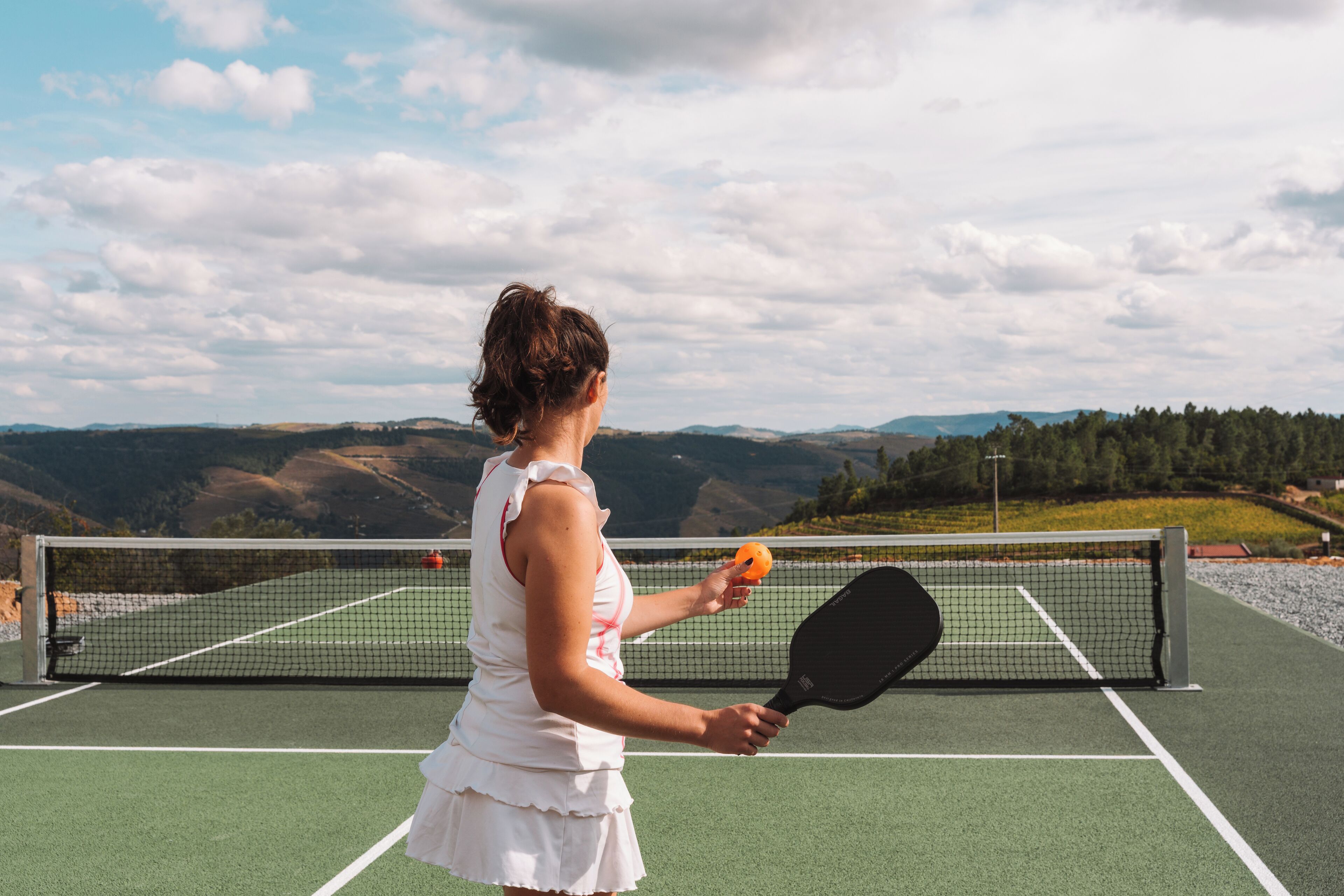A tennis court with a net, surrounded by green fields and distant hills.