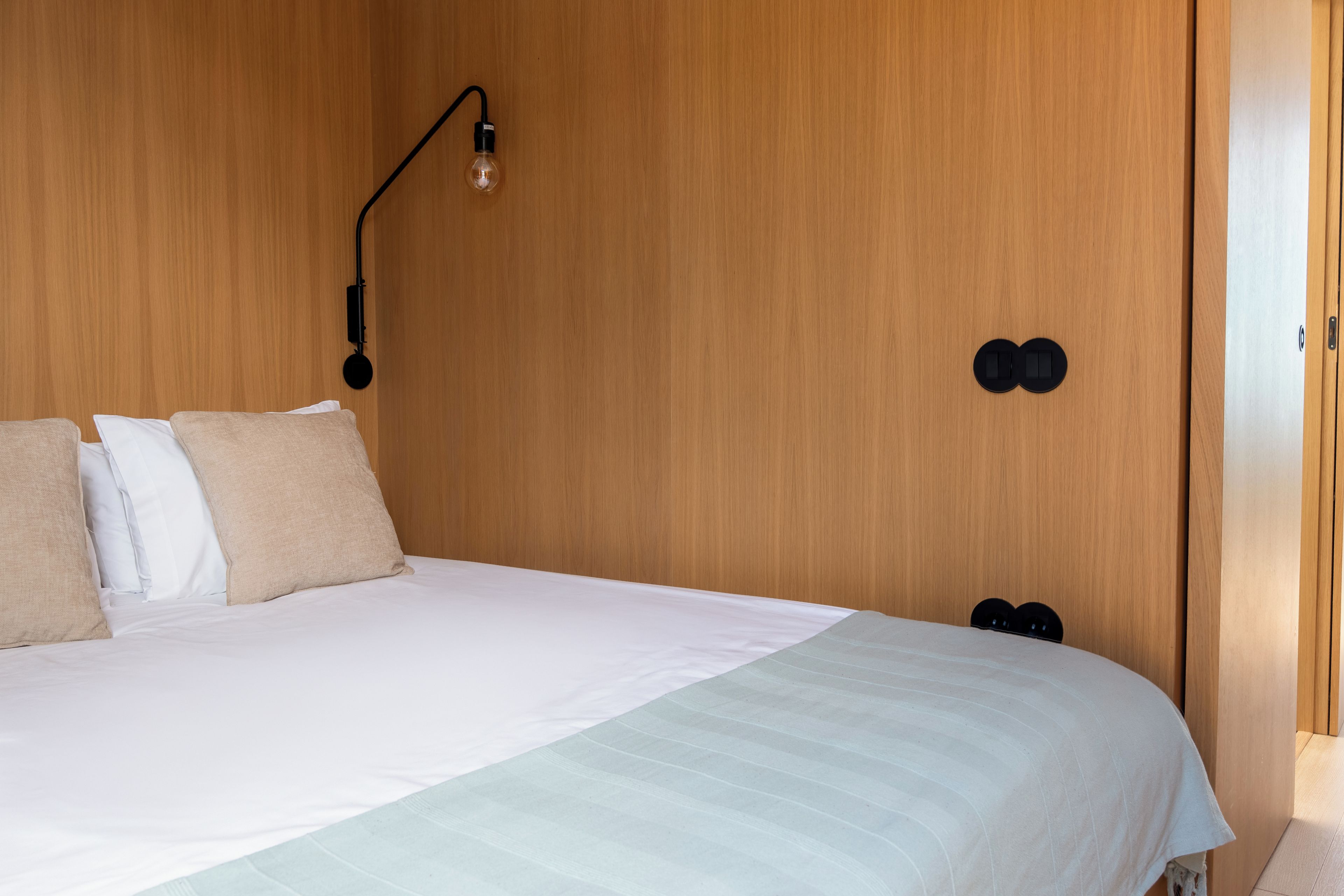 Een slaapkamer met een houten hoofdbord, een bed met wit beddengoed, een wandlamp en een deur met een hendel.