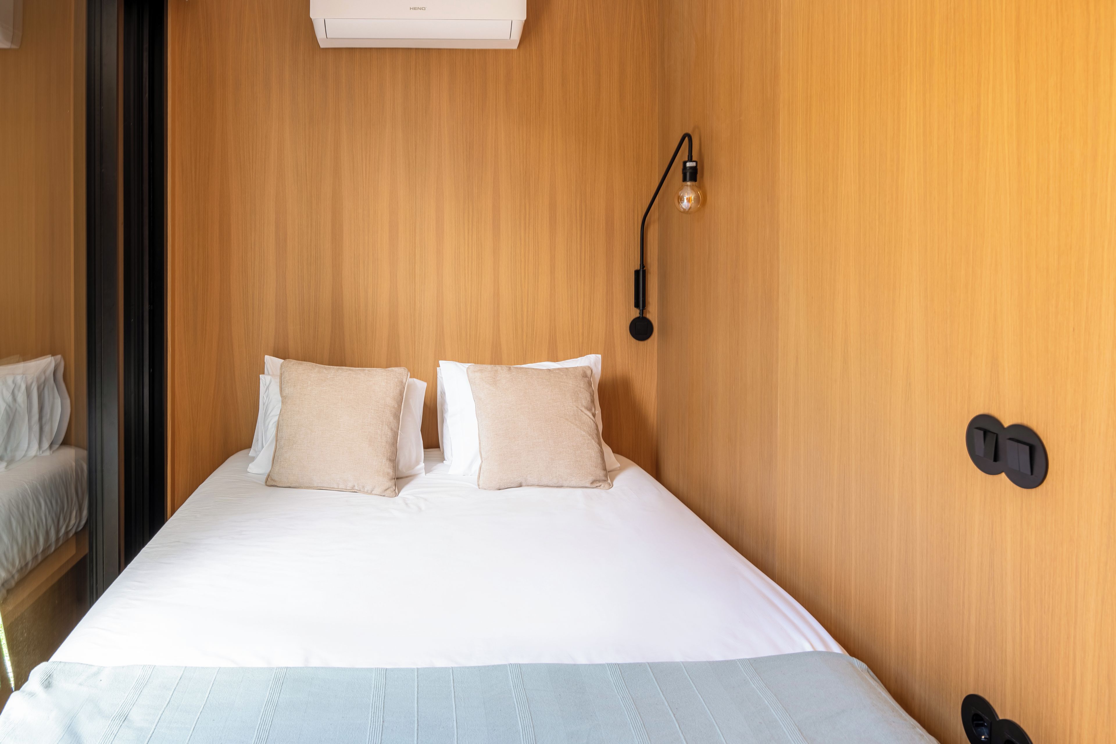 Una habitación de hotel con pared de madera, una cama con ropa de cama blanca, dos almohadas color beige, una lámpara y un enchufe de pared.