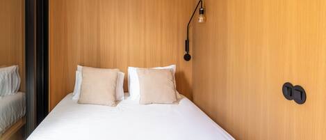 Una habitación de hotel con pared de madera, una cama con ropa de cama blanca, dos almohadas color beige, una lámpara y un enchufe de pared.