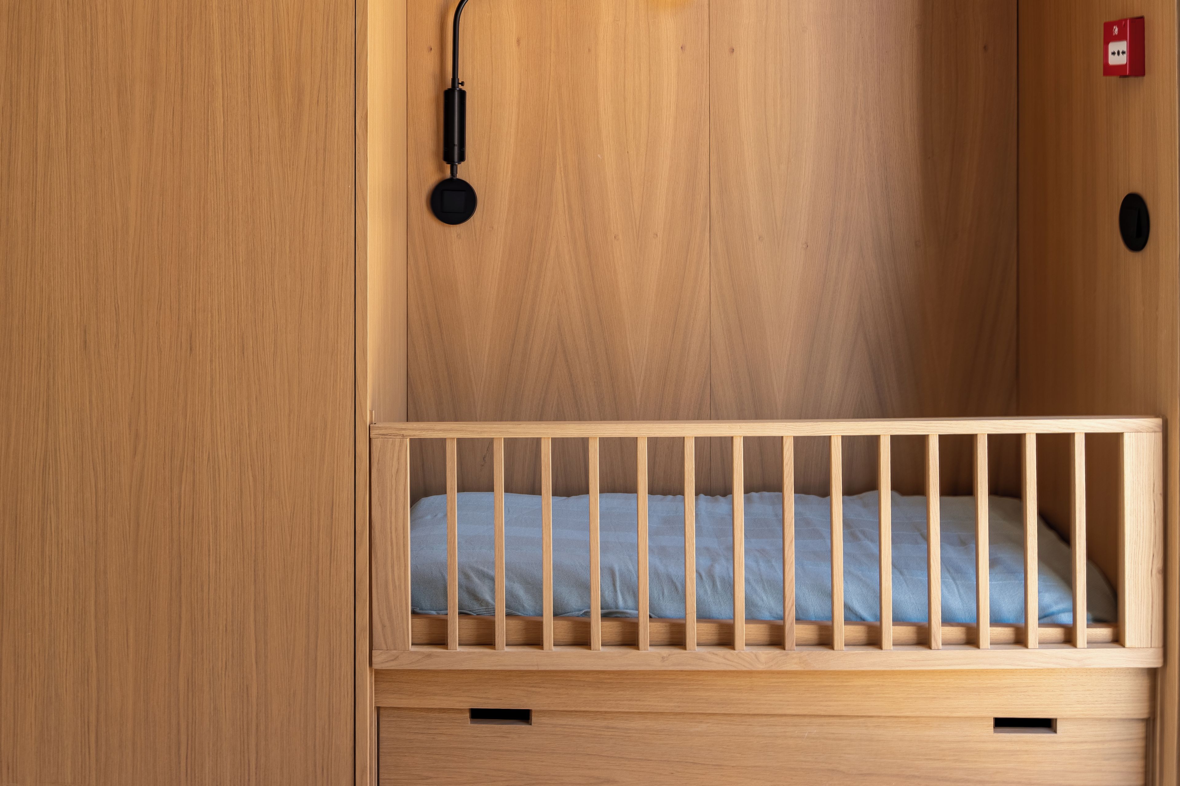 A wooden crib with a blue mattress, a small drawer below, and a red emergency light on the wall.