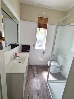 A bathroom with a white sink, mirror, and toilet, wooden flooring, and a window with blinds.