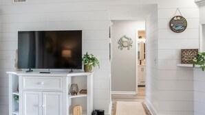 Un salon moderne avec un téléviseur à écran plat posé sur une console blanche, une plante en pot et un panneau décoratif mural.
