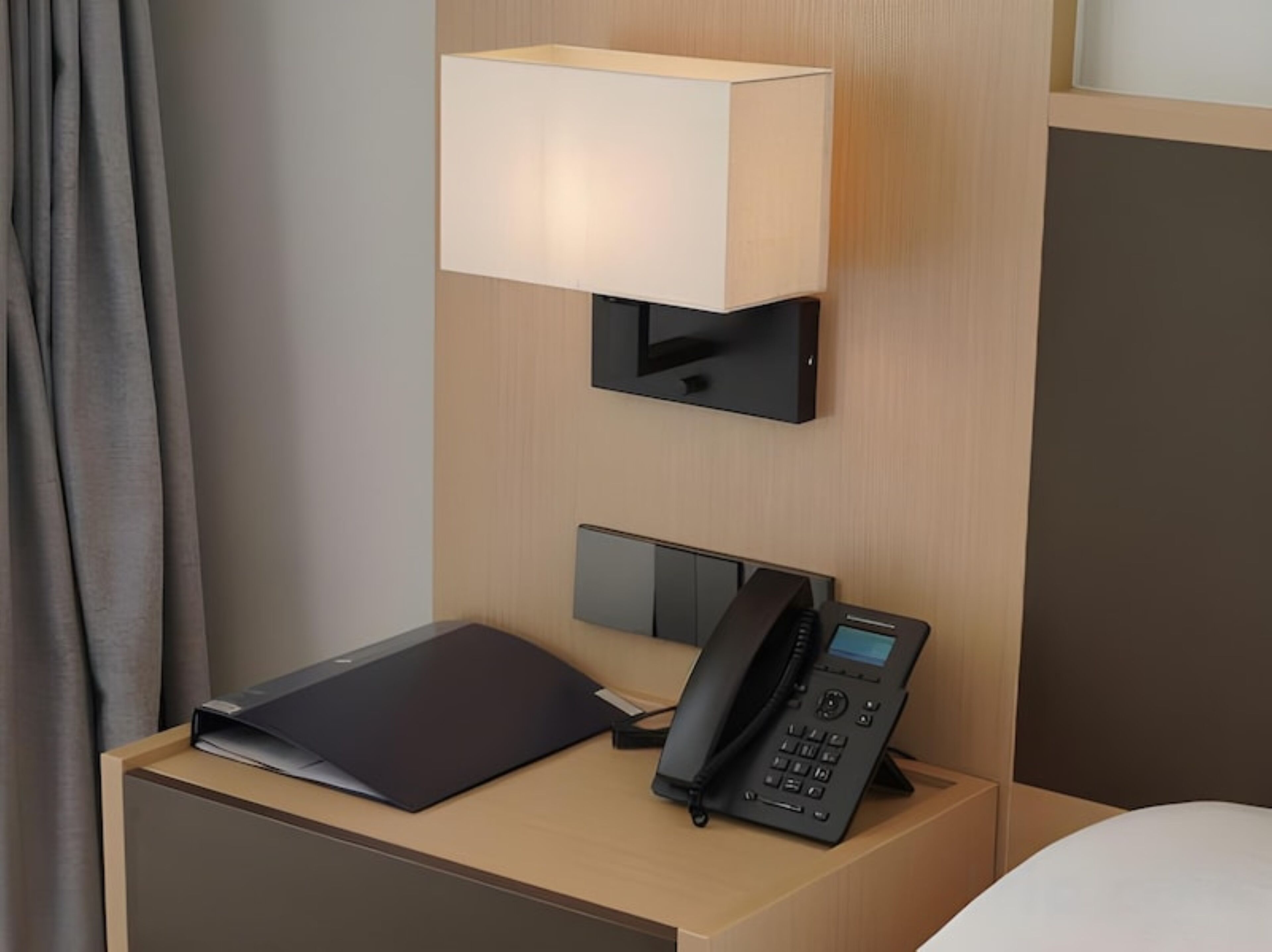 A hotel room with a bedside table, a lamp, a telephone, and a black folder.