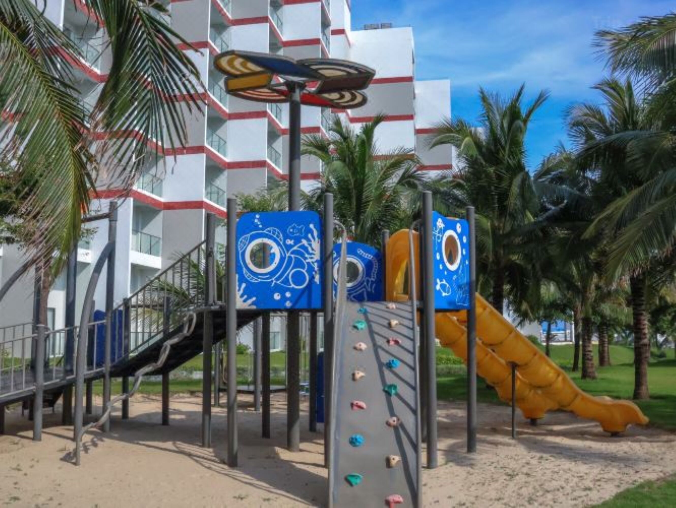A playground with climbing structures and a slide, surrounded by palm trees and a building with balconies.
