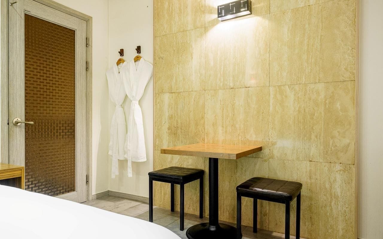 A bathroom with a wooden table and two stools, a white robe hanging on the wall, and a door with a brick pattern.