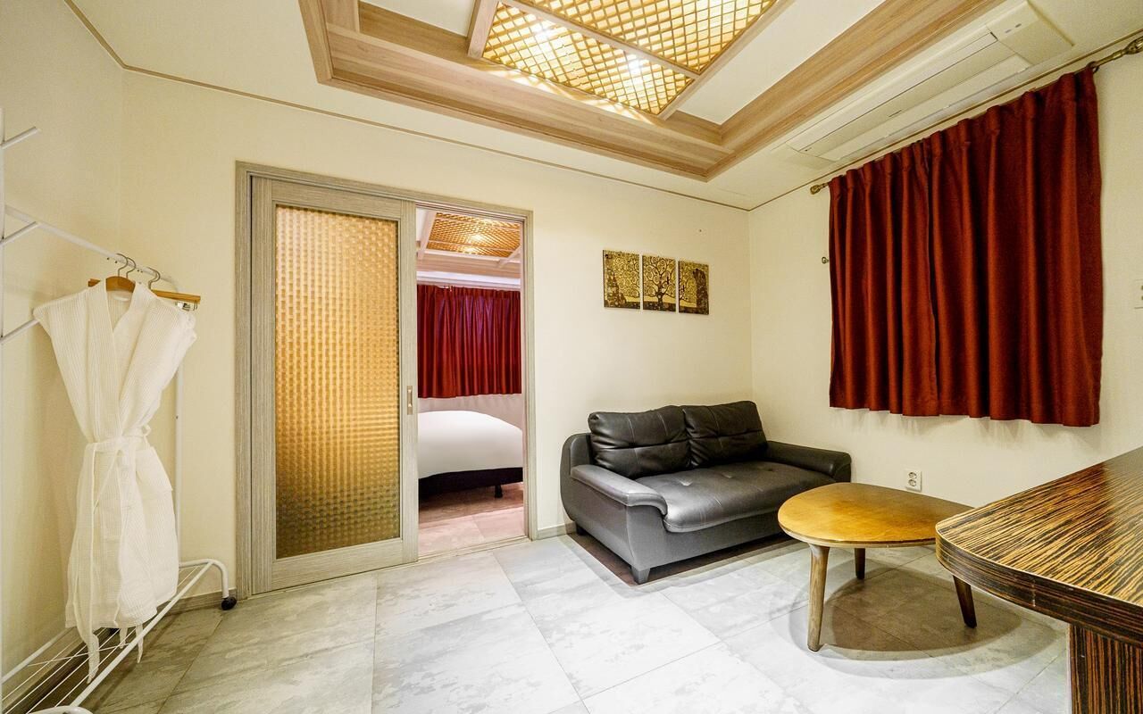 Habitación de hotel con sofá, una mesita redonda, perchero y una puerta corrediza que da a un dormitorio.