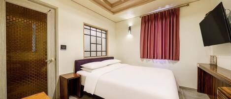A hotel room with a bed, a TV, a window with a curtain, a wooden table, and a door with a textured glass panel.