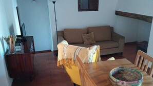 A living room with a sofa, a dining table with chairs, a lamp, and a shelf with decorative items.