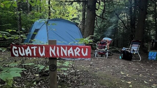 Une tente portant l’inscription « GUATU NINARU » est installée dans une zone boisée, avec une chaise et un chariot à proximité.
