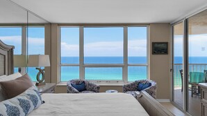 Une chambre d’hôtel avec un grand lit, deux fauteuils, une petite table et une vue sur l’océan grâce à de grandes fenêtres.