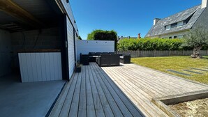 A wooden deck with outdoor furniture, a white wall, and a hedge.