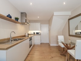 A modern kitchen with a wooden countertop, stainless steel sink, and white cabinets. There is a microwave, toaster, and coffee maker on the countertop. A glass table with two white chairs is in the dining area.