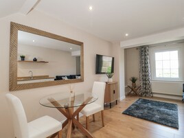 A modern living room with a glass-top dining table, white chairs, a large mirror, a TV, and a window with curtains.