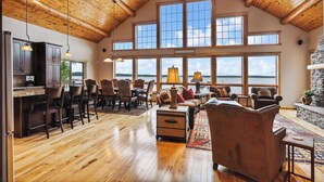 A spacious living area with wooden flooring, a stone fireplace, and large windows offering a view of the outdoors.