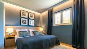 A bedroom with a large bed, two bedside tables with lamps, a window with curtains, and framed pictures on the wall.