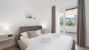 A neatly made bed with white linens and two pillows, a bedside table with a lamp, a wall-mounted artwork, and a view of buildings through a window with curtains.