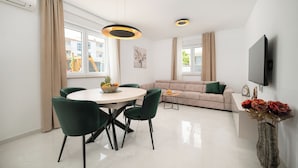A modern living room with a sofa, a dining table with chairs, a flat-screen TV mounted on the wall, and a vase with flowers on a side table.