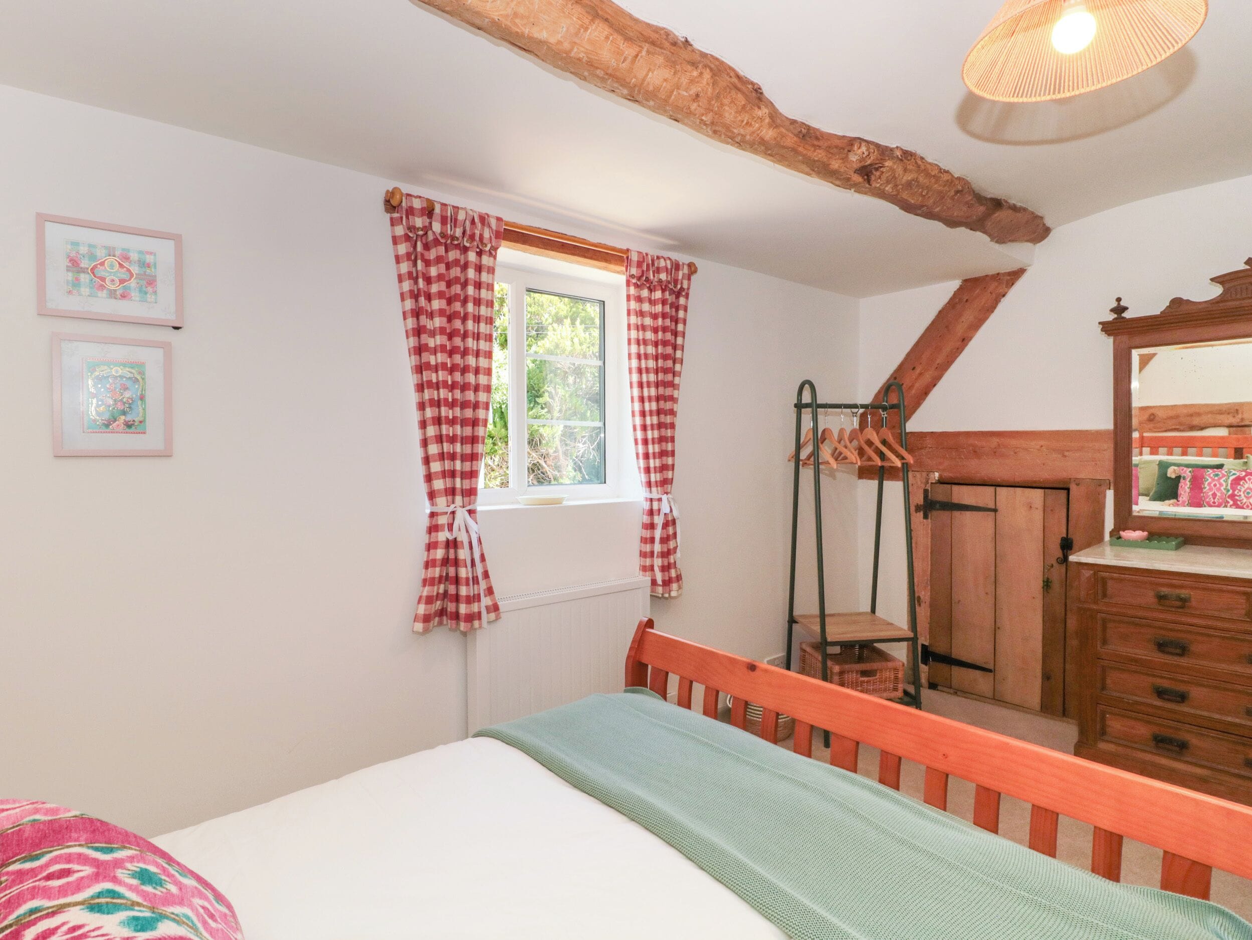 A bedroom with a bed, a window with checkered curtains, a wooden dresser, and a mirror.