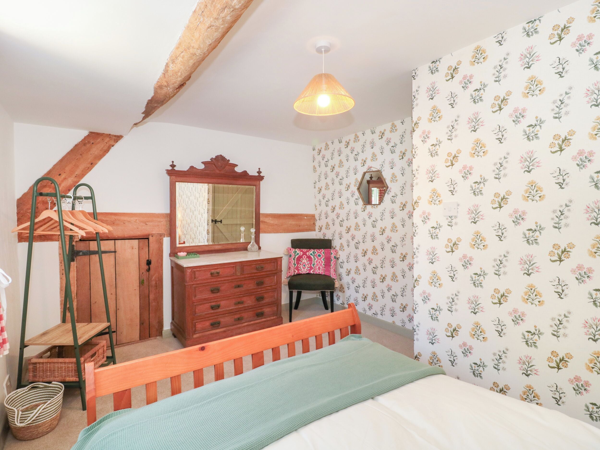Ein Schlafzimmer mit einem hölzernen Bett, einem Schminktisch, einem Spiegel, einem Kleiderständer und floralem Wandpapier.