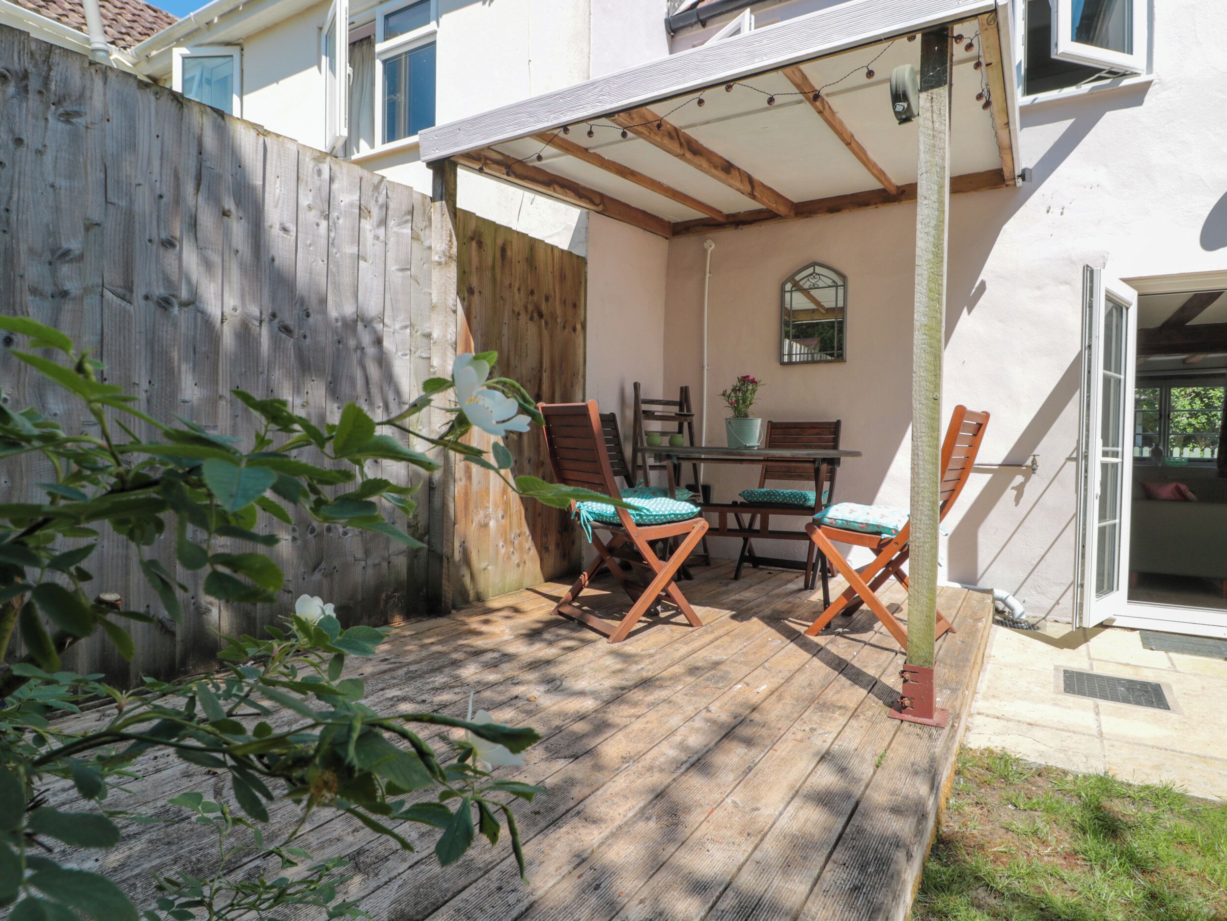 Una terraza de madera con una mesa y sillas, una cerca de madera, y un edificio con ventana y puerta.