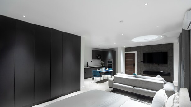 A modern bedroom with a large bed, a sofa, and a dining area with a TV and kitchen in the background. - The VVS Apartments - Studio Apartment with Balcony (Accra)