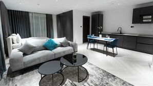 A modern living room with a grey sofa, a coffee table, and a dining area with blue chairs. - The VVS Apartments - Studio Apartment with Balcony (Accra)