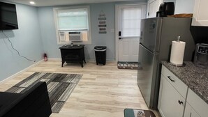 Cocina compacta con refrigerador, estufa y microondas. Hay dos ventanas con persianas, una mesita negra y un cesto de basura.
