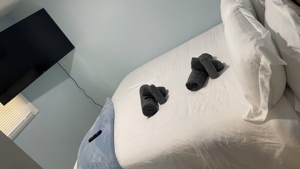 Un dormitorio con cama, almohadas, un control remoto y un televisor montado en la pared.