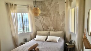 A bedroom with a bed, two pillows, a rolled towel, a bedside table with a lamp, a small table with a clock, and a window with curtains. - Villa Almazara (Xalo)