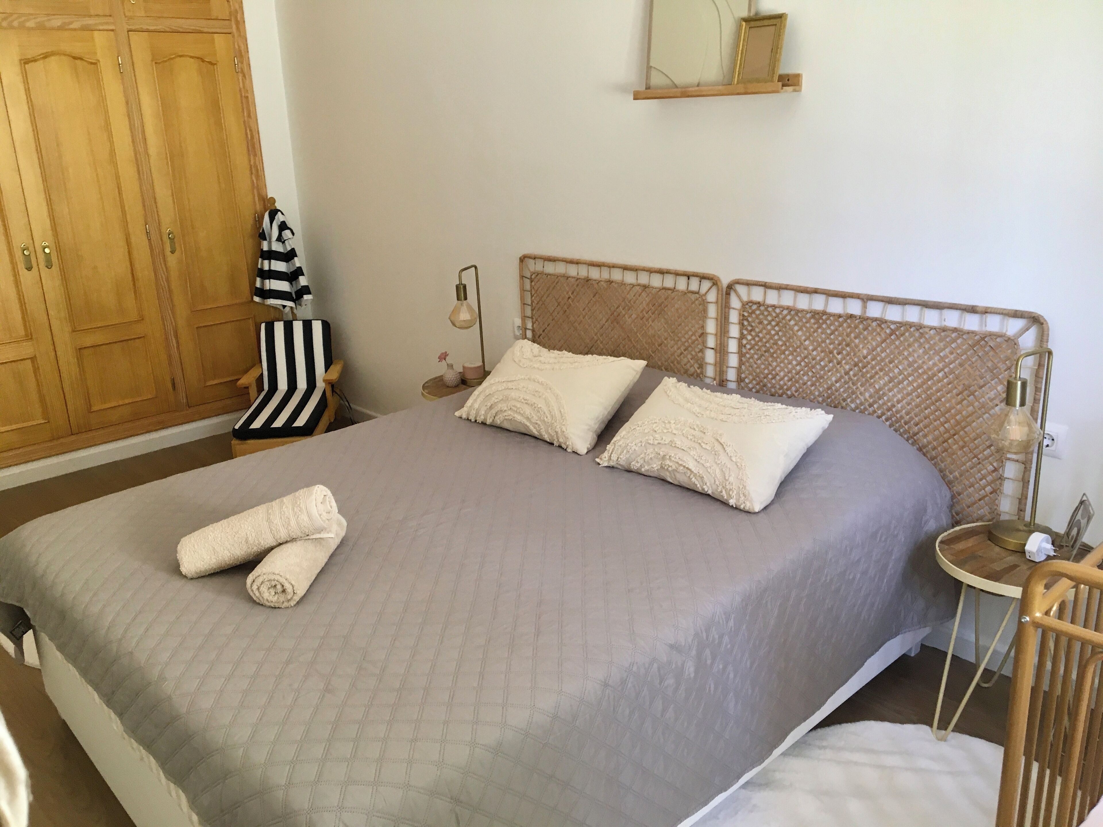 Ein ordentlich bezogenes Bett mit einer gesteppten, grauen Tagesdecke und zwei Zierkissen, ein Holzschrank, ein gestreifter Stuhl und ein Nachttisch mit Lampe und kleiner Vase.