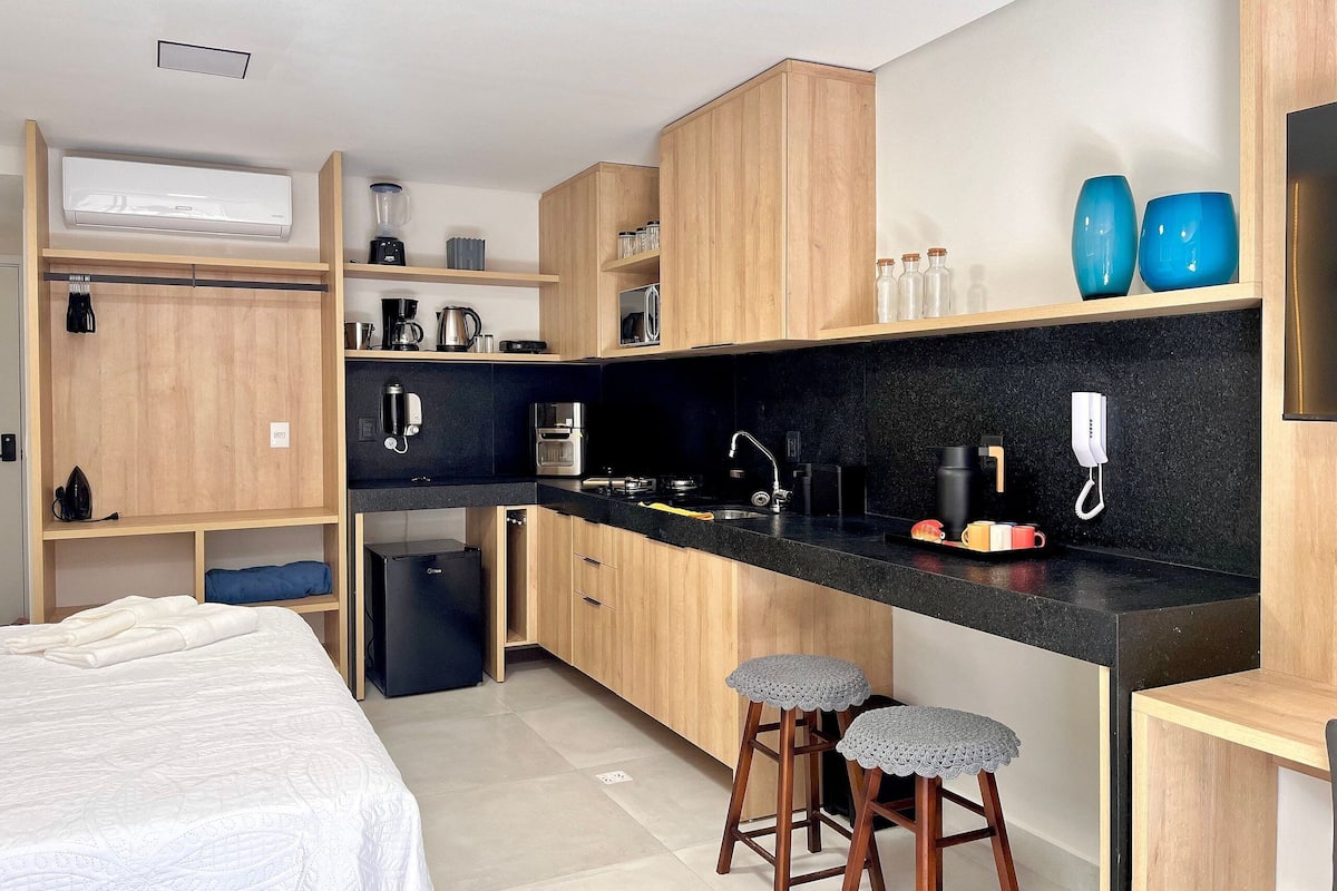 A compact kitchen with wooden cabinets, a black countertop, and a small refrigerator. There are two stools and a coffee machine.