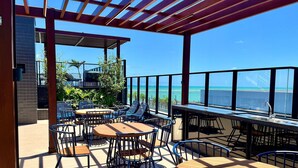 An outdoor patio with tables and chairs, a pergola, and a view of the ocean. - Hyde Park Select - by Living (Joao Pessoa)