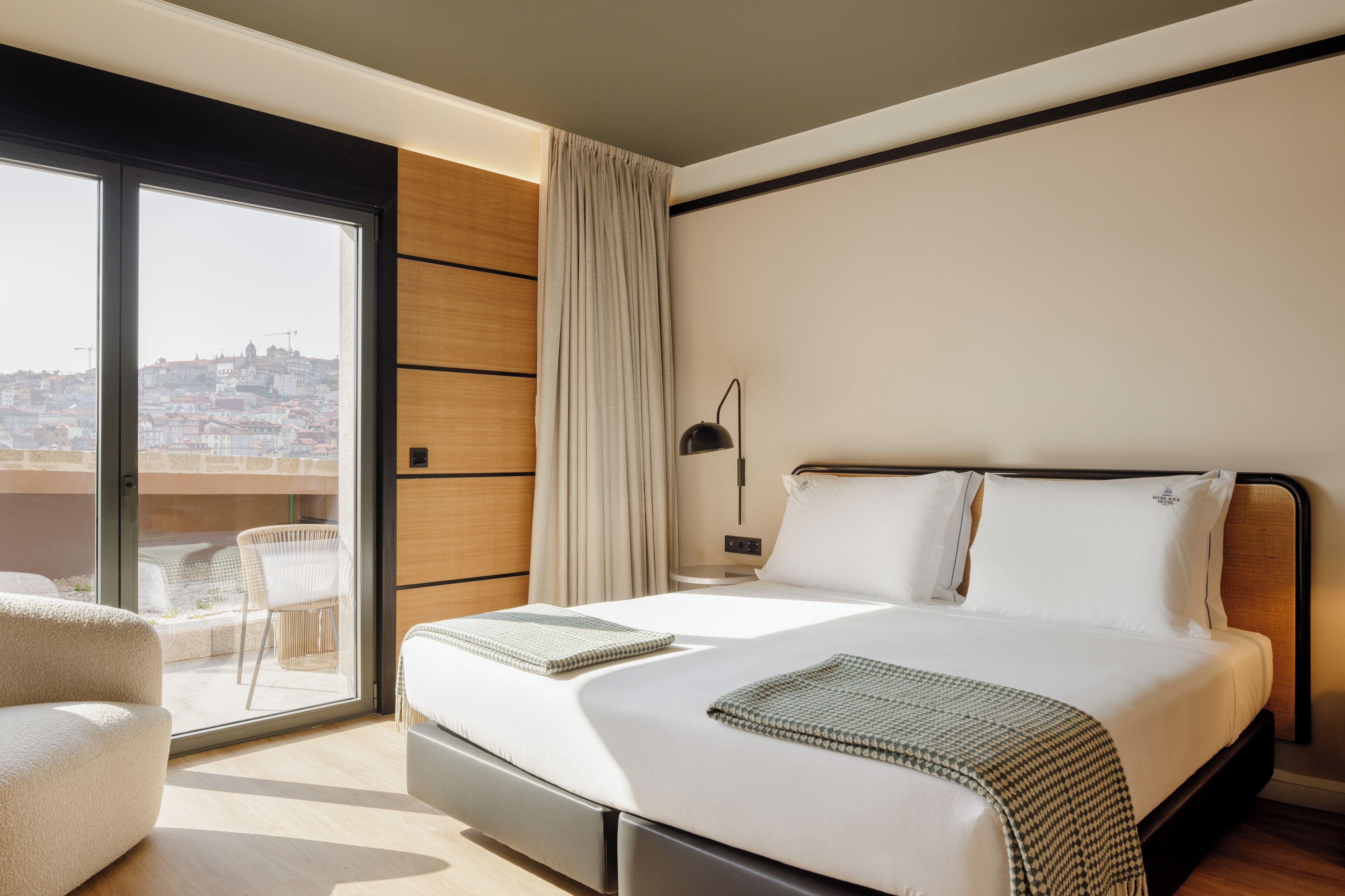 A modern hotel room with a large bed, a view of the city through a sliding glass door, and a balcony with a chair.