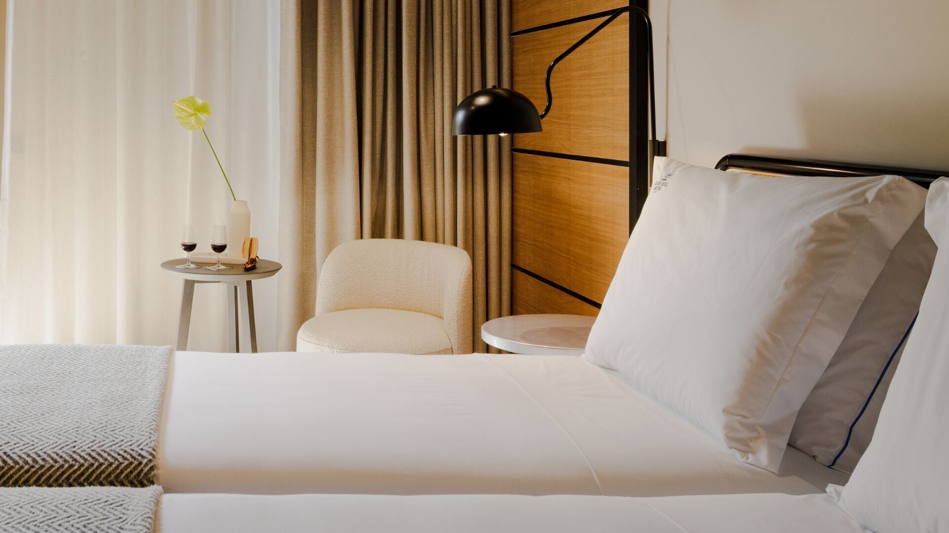 A hotel room with a bed, a chair, a small table with a vase and glasses, and a lamp.